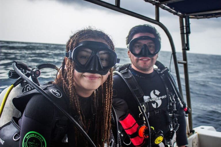 Two scuba divers in Scubapro wetsuits on a boat with diving gear, ocean waves and cloudy sky in the background, featured in the blog post ‘Scuba Diving Travels & Expeditions to Costa Rica and Abroad.’