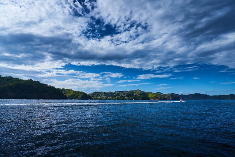 Scenic coastal view with a boat crossing deep blue water, green hills and buildings in the background under a dramatic cloudy sky, featured in the blog post ‘Scuba Diving Trips & Tours in Guanacaste Costa Rica.’
